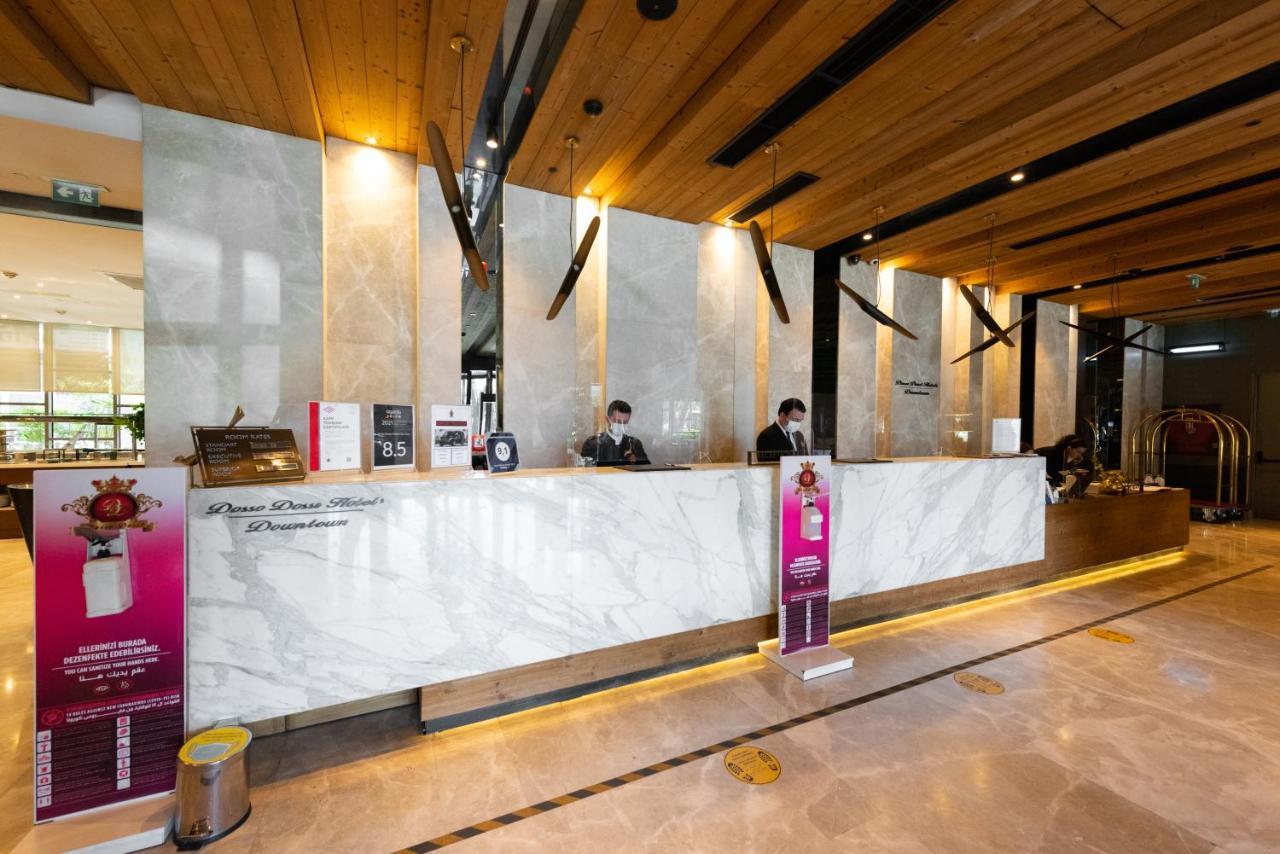 Dosso Dossi Hotels & Spa Downtown Istanbul Luaran gambar The photo shows a hotel reception area. The front desk is made of elegant marble, and the space features wooden accents on the ceiling. Behind the counter, two staff members are visible, likely assisting guests. There are signs and promotional materi