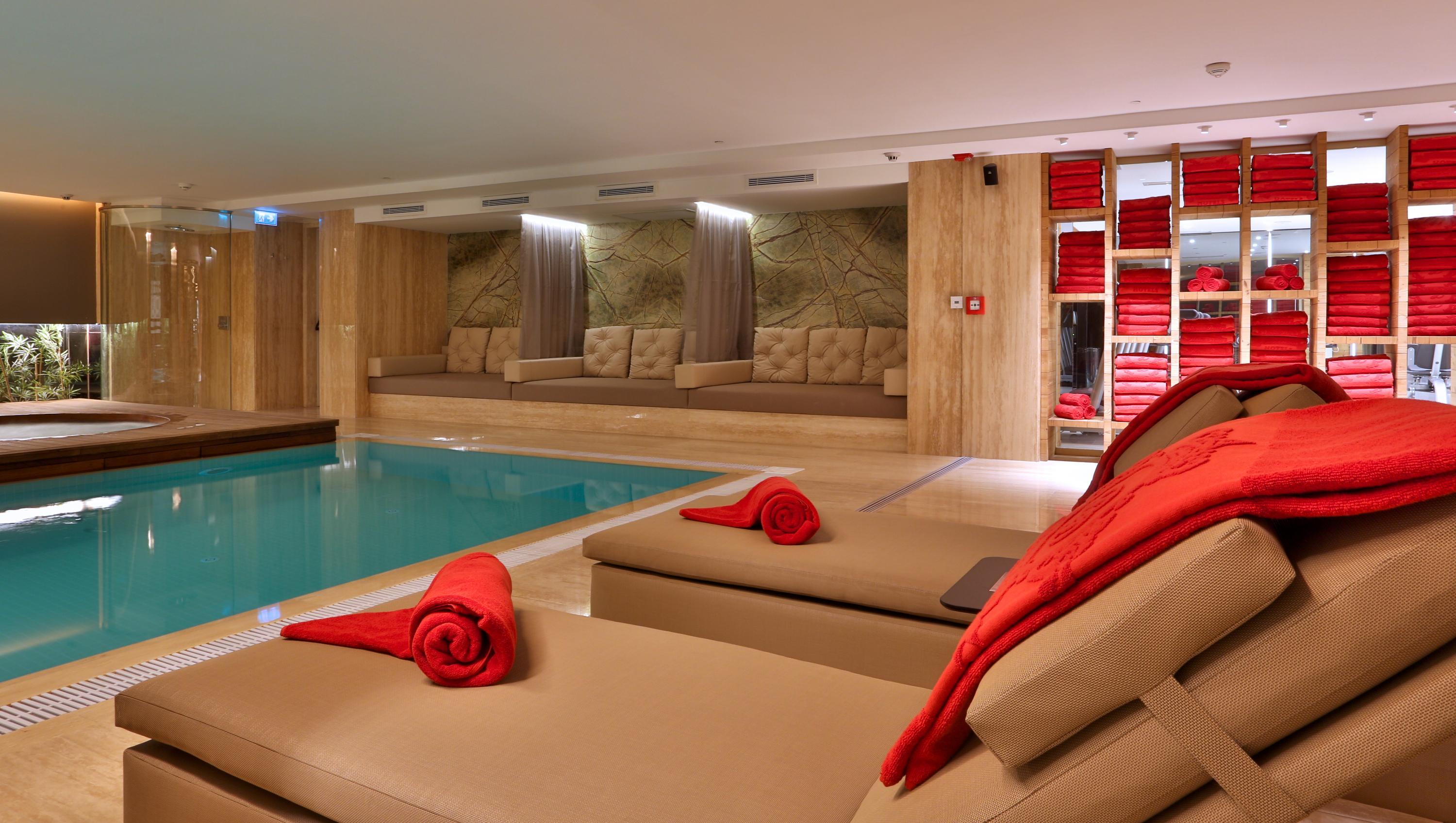 Dosso Dossi Hotels & Spa Downtown Istanbul Luaran gambar The photo shows a modern, luxurious spa area featuring a swimming pool. In the foreground, there are comfortable lounge chairs with red towels rolled and placed on them. The walls are decorated with wood paneling, and there are several plush seating 